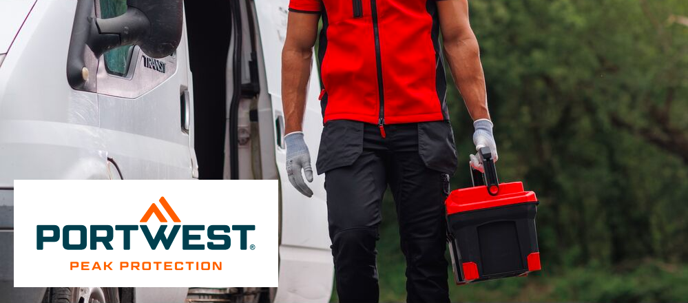 Man sieht den Bereich zwischen Schultern und Waden einer Person in Arbeitskleidung. Der Mann trägt eine rote Softshellweste, ein rotes T-shirt darunter, eine schwarze Hose mit Taschen und weiße Handschuhe. Er trägt einen Werkzeugkoffer in seiner linken Hand. Links neben ihm im Hintergrund sieht man einen weißen Transporter, der restliche Hintergrund ist von grünen Laubbäumen bedeckt. Das blau-orange Logo des Herstellers Portwest auf weißem Grund befindet sich im linken unteren Bildrand. 