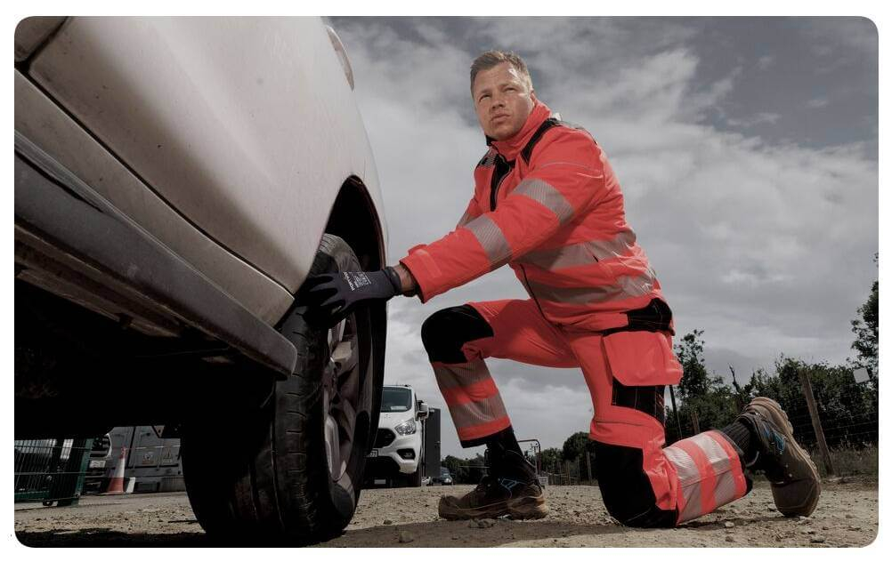 Ein Mann in oranger Warnschutzkleidung kniet vor einem weißen Auto und macht etwas am Reifen. Im Hintergrund sieht man einen bewölkten Himmel. Ein Link zu unserer Knieschutz-Kollektion ist hinterlegt.