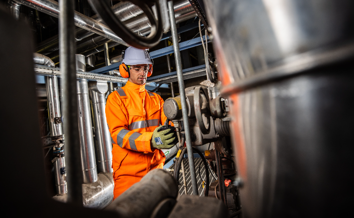 Arbeiter mit weißem Helm und Ohrenschutz in oranger Arbeitskleidung mit Handschuhen vor einem industriellen Hintergrund. Das Bild ist mit der Kollektion an Arbeitshandschuhen verlinkt. 