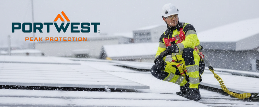 Ein Arbeiter in einem gelben Warnschutzoverall mit Reflexstreifen kniet auf einem verschneiten Dach neben einer Photovoltaikanlage. Er tr&auml;gt einen wei&szlig;en Schutzhelm und eine Absturzsicherung. Im Hintergrund sind weitere schneebedeckte D&auml;cher zu sehen und im linken, oberen Bildbereich findet sich das Logo des Herstellers Portwest in Blau und Orange.