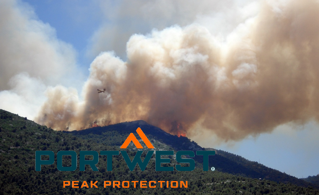 Ein Waldbrand tobt auf einem grünen Berghang, dichte Rauchwolken steigen in den Himmel. Ein Löschflugzeug ist im Anflug und wirft Wasser oder Löschmittel über die Flammen. Im Vordergrund ist das Logo der Marke Portwest zu sehen – darunter der Slogan „Peak Protection“. Ein Link zu unserer Auswahl an Feuerwehrkleidung ist hinterlegt.