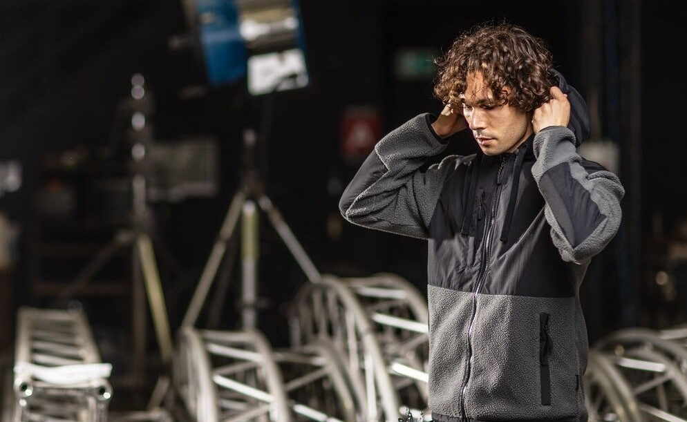 Mann mit lockigem Haar trägt eine graue Fleecejacke mit Kapuze und schwarzen Einsätzen, steht in einem industriellen Umfeld mit Traversen und Scheinwerfern. Ein Link zu unserer Fleece-Kollektion ist hinterlegt.