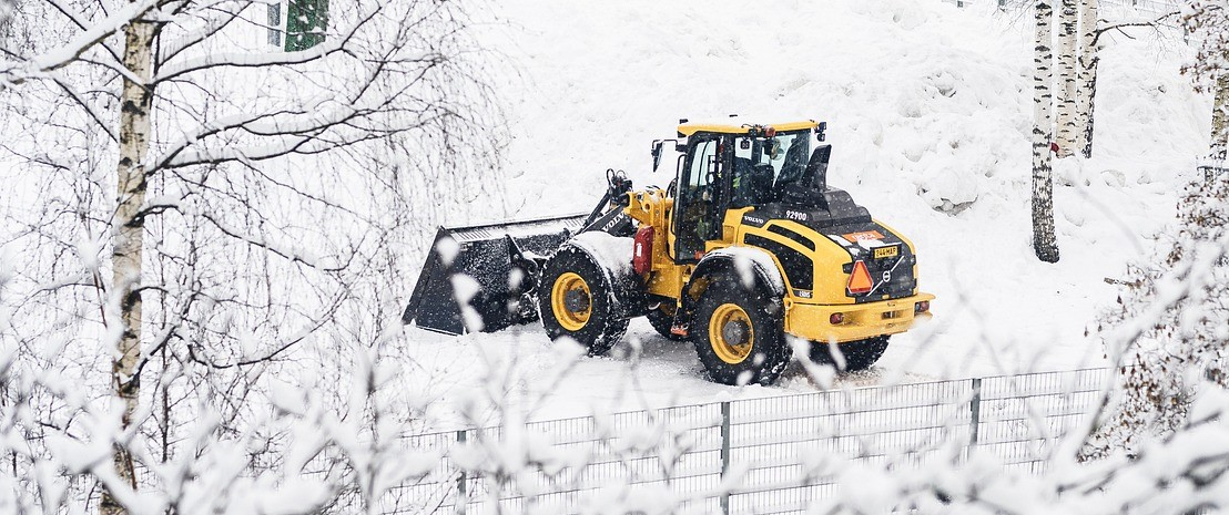 Gelber Traktor mit Schaufel in verschneiter Landschaft mit Zäunen und Birken.