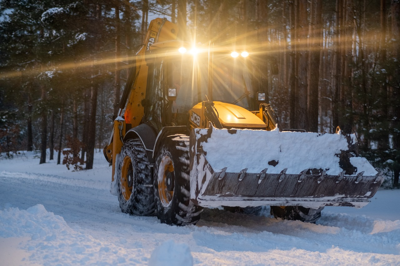 Gelber Traktor mit großer Schaufel auf einem verschneiten Waldweg. Die Dämmerung bricht langsam ein und das Fahrzeug hat ein Licht an.