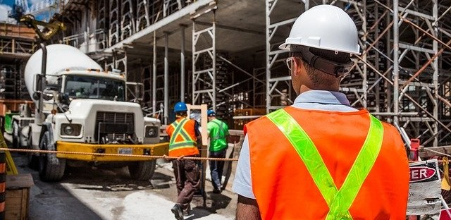 Rückenansicht eines Bauarbeiters mit Warnweste, Schutzhelm und Schutzbrille auf einer Baustelle mit Gerüsten.