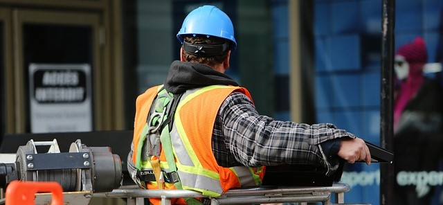 Rückenansicht eines Bauarbeiters mit Schutzhelm und Absturzsicherung auf einer Hebebühne.